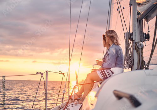 Female friends relaxing on the yacht with glasses of wine in the hands, during sunset on the high seas.