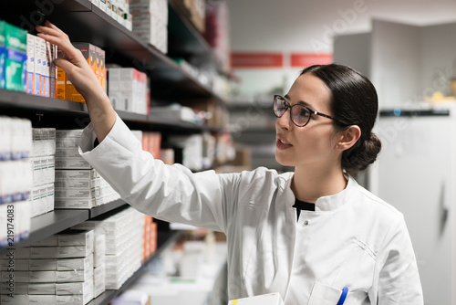 Portrait of a dedicated female pharmacist taking a medicine from the shelf, while wearing eyeglasses and lab coat during work in a modern drugstore with various pharmaceutical products photo