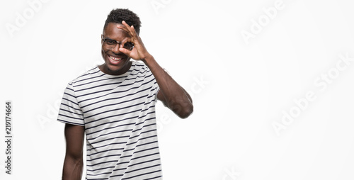 Young african american man wearing glasses and navy t-shirt doing ok gesture with hand smiling, eye looking through fingers with happy face.