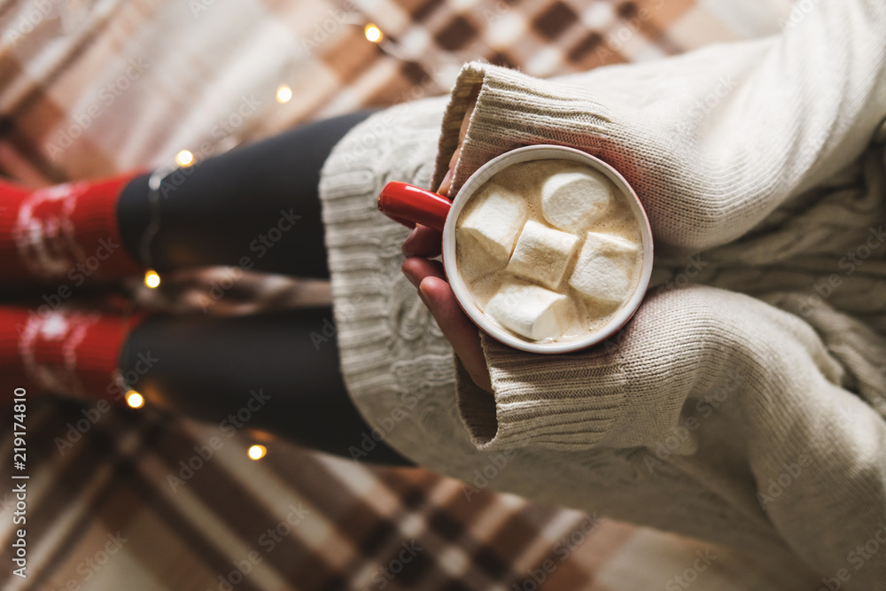 Women's hands and feet in sweater and woolen cozy red socks holding cup of hot coffee with marshmallow, sitting on plaid with garland, tablet and notebook. Concept winter comfort, morning drinking. Women's hands and feet in sweater and woolen cozy red socks holding cup of hot coffee with marshmallow, sitting on plaid with garland, tablet and notebook. Concept winter comfort, morning drinking.