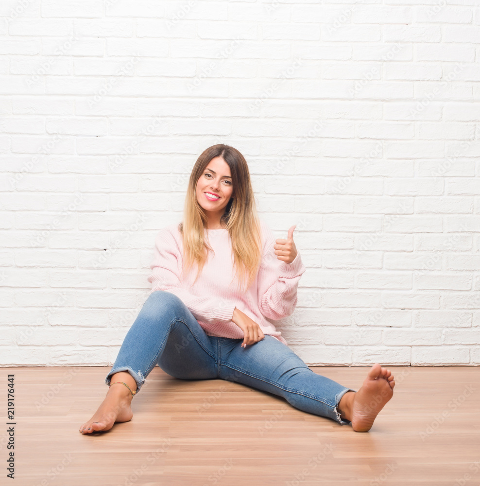 Young adult woman sitting on the floor over white brick wall doing happy thumbs up gesture with hand. Approving expression looking at the camera with showing success. Young adult woman sitting on the floor over white brick wall doing happy thumbs up gesture with hand. Approving expression looking at the camera with showing success.