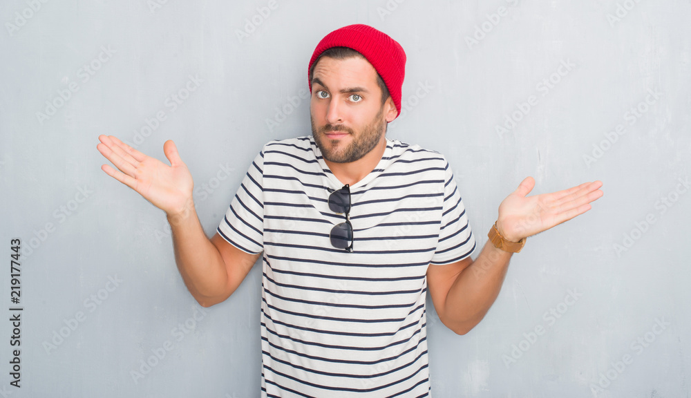 Naklejka premium Handsome young hipster man over grey grunge wall wearing navy t-shirt and wool cap clueless and confused expression with arms and hands raised. Doubt concept.