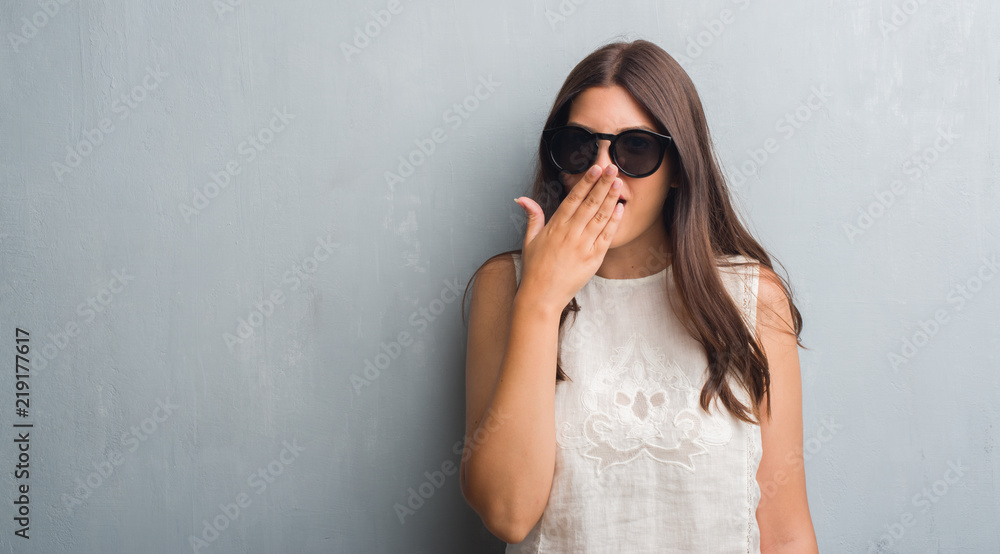 Young brunette woman over grunge grey wall wearing fashion sunglasses cover mouth with hand shocked with shame for mistake, expression of fear, scared in silence, secret concept Young brunette woman over grunge grey wall wearing fashion sunglasses cover mouth with hand shocked with shame for mistake, expression of fear, scared in silence, secret concept