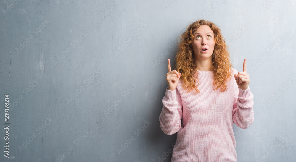 Young redhead woman over grey grunge wall wearing pink sweater amazed and surprised looking up and pointing with fingers and raised arms. Young redhead woman over grey grunge wall wearing pink sweater amazed and surprised looking up and pointing with fingers and raised arms.