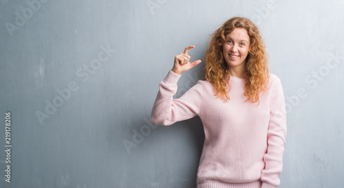 Young redhead woman over grey grunge wall wearing pink sweater smiling and confident gesturing with hand doing size sign with fingers while looking and the camera. Measure concept.