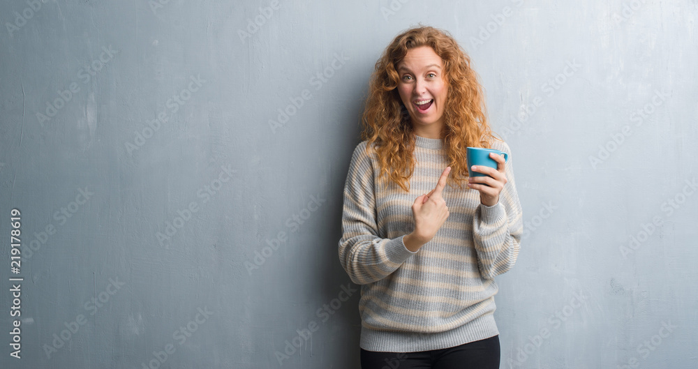 Young redhead woman over grey grunge wall drinking a cup of coffee very happy pointing with hand and finger Young redhead woman over grey grunge wall drinking a cup of coffee very happy pointing with hand and finger
