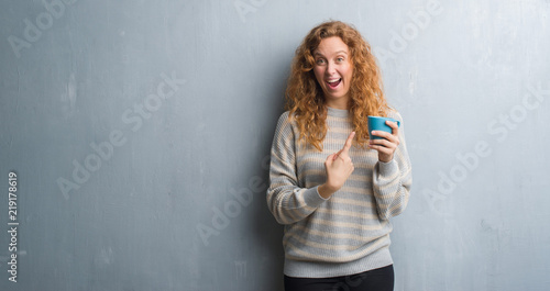 Young redhead woman over grey grunge wall drinking a cup of coffee very happy pointing with hand and finger