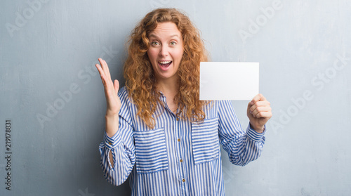 Young redhead woman over grey grunge wall holding blank card very happy and excited, winner expression celebrating victory screaming with big smile and raised hands