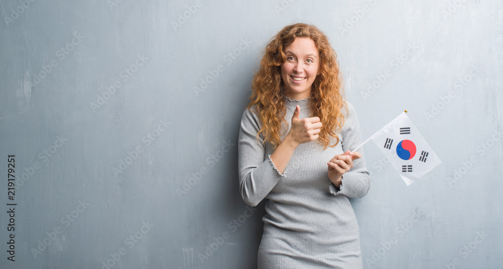 Young redhead woman over grey grunge wall holding flag of South Korea happy with big smile doing ok sign, thumb up with fingers, excellent sign Young redhead woman over grey grunge wall holding flag of South Korea happy with big smile doing ok sign, thumb up with fingers, excellent sign