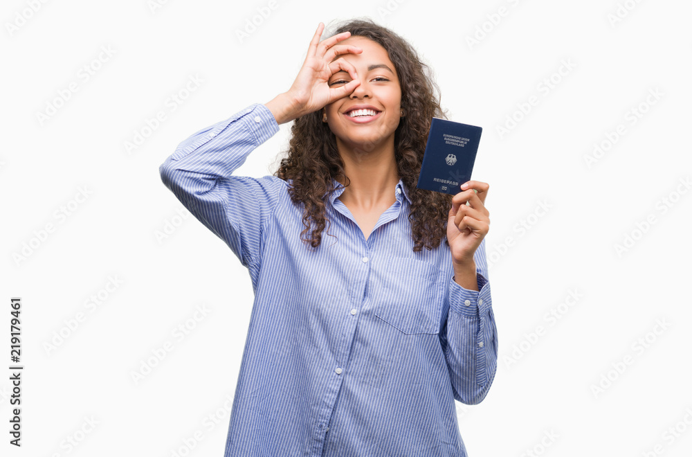 Young hispanic woman holding passport of Germany with happy face smiling doing ok sign with hand on eye looking through fingers Young hispanic woman holding passport of Germany with happy face smiling doing ok sign with hand on eye looking through fingers
