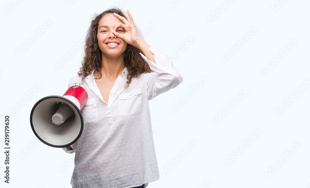 Young hispanic woman holding megaphone with happy face smiling doing ok sign with hand on eye looking through fingers Young hispanic woman holding megaphone with happy face smiling doing ok sign with hand on eye looking through fingers