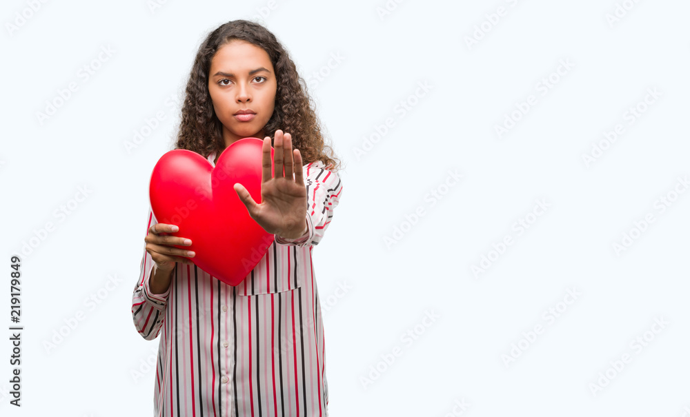 Young hispanic woman in love holding red heart with open hand doing stop sign with serious and confident expression, defense gesture Young hispanic woman in love holding red heart with open hand doing stop sign with serious and confident expression, defense gesture
