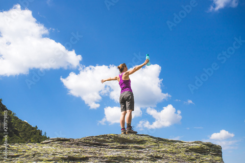 Canvastavla Junge Frau in Wanderschuhen steht auf Felsbrocken, blauer Himmel im Hintergrund