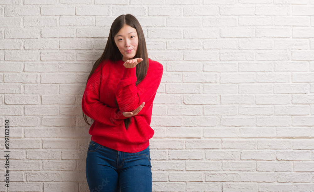 Young Chinese woman over brick wall looking at the camera blowing a kiss with hand on air being lovely and sexy. Love expression. Young Chinese woman over brick wall looking at the camera blowing a kiss with hand on air being lovely and sexy. Love expression.
