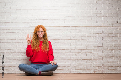 Young redhead woman sitting over brick wall showing and pointing up with fingers number four while smiling confident and happy. © Krakenimages.com
