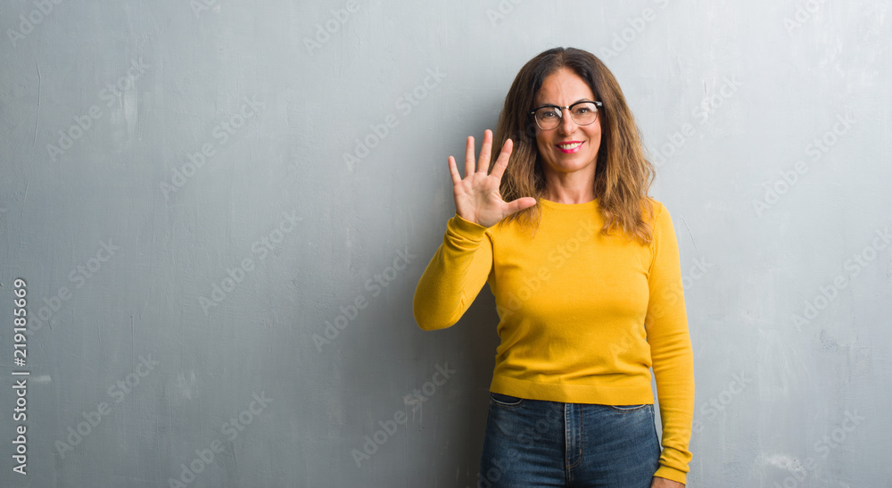 Middle age hispanic woman over grey wall wearing glasses showing and pointing up with fingers number five while smiling confident and happy. Middle age hispanic woman over grey wall wearing glasses showing and pointing up with fingers number five while smiling confident and happy.