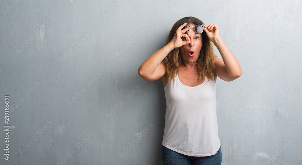 Middle age hispanic woman over grey wall wearing sunglasses doing ok gesture shocked with surprised face, eye looking through fingers. Unbelieving expression. Middle age hispanic woman over grey wall wearing sunglasses doing ok gesture shocked with surprised face, eye looking through fingers. Unbelieving expression.