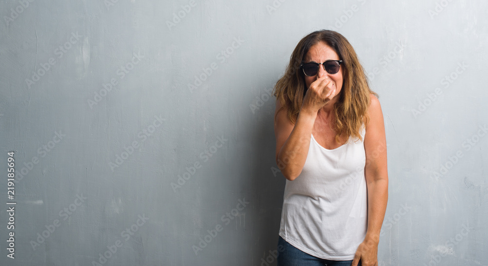 Middle age hispanic woman over grey wall wearing sunglasses smelling something stinky and disgusting, intolerable smell, holding breath with fingers on nose. Bad smells concept. Middle age hispanic woman over grey wall wearing sunglasses smelling something stinky and disgusting, intolerable smell, holding breath with fingers on nose. Bad smells concept.