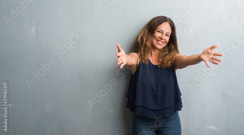 Middle age hispanic woman standing over grey grunge wall looking at the camera smiling with open arms for hug. Cheerful expression embracing happiness.