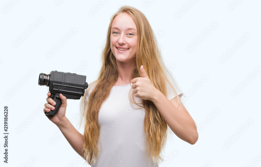Blonde woman teenager filming holding super 8 video camera happy with big smile doing ok sign, thumb up with fingers, excellent sign Blonde woman teenager filming holding super 8 video camera happy with big smile doing ok sign, thumb up with fingers, excellent sign