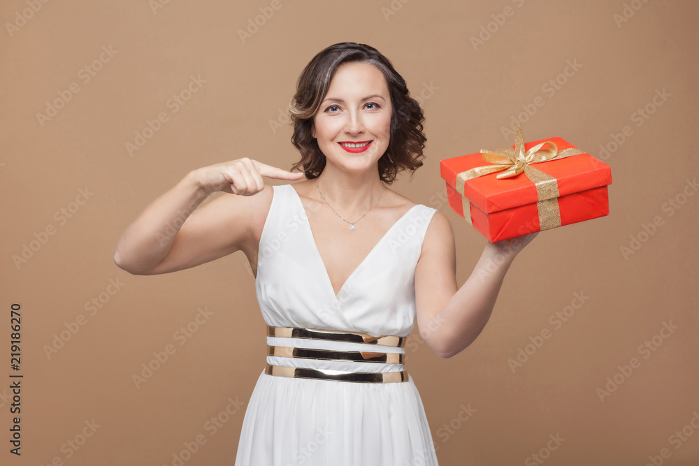 Toothy smile elegant brunette woman in white dress pointing at red gift box and looking at camera with happy face. Emotion and feeling concept. Studio shot, indoor, isolated on light brown background Toothy smile elegant brunette woman in white dress pointing at red gift box and looking at camera with happy face. Emotion and feeling concept. Studio shot, indoor, isolated on light brown background