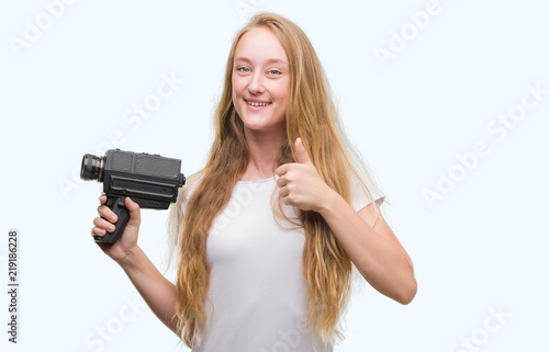 Blonde woman teenager filming holding super 8 video camera happy with big smile doing ok sign, thumb up with fingers, excellent sign