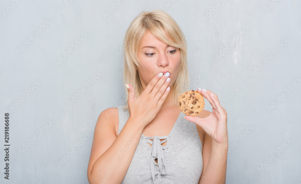 Caucasian adult woman over grey grunge wall eating chocolate cooky cover mouth with hand shocked with shame for mistake, expression of fear, scared in silence, secret concept Caucasian adult woman over grey grunge wall eating chocolate cooky cover mouth with hand shocked with shame for mistake, expression of fear, scared in silence, secret concept