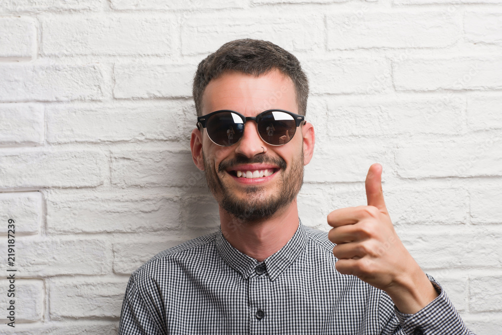 Naklejka premium Young adult man wearing sunglasses standing over white brick wall happy with big smile doing ok sign, thumb up with fingers, excellent sign