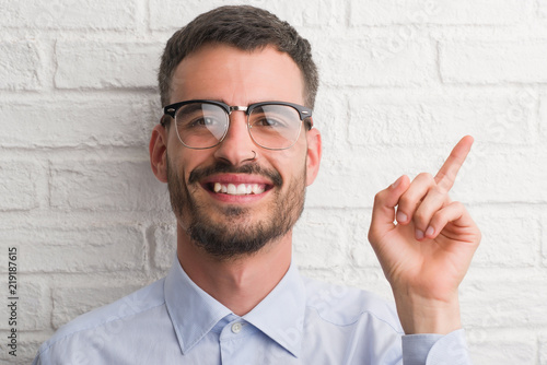 Young adult business man standing over white brick wall very happy pointing with hand and finger to the side