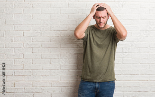 Young caucasian man standing over white brick wall suffering from headache desperate and stressed because pain and migraine. Hands on head.