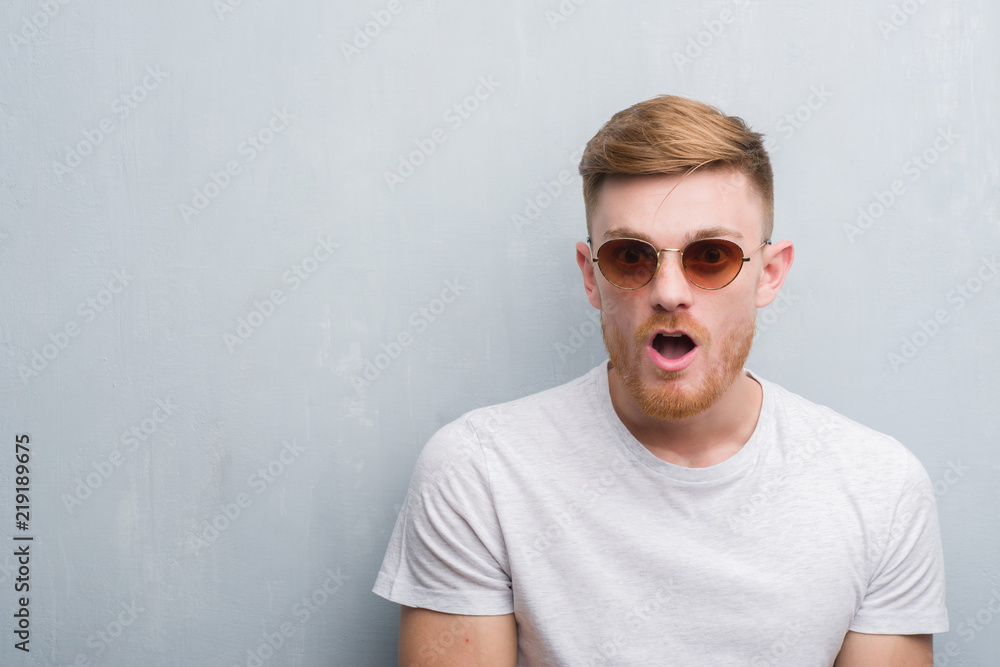 Young redhead man over grey grunge wall wearing retro sunglasses scared in shock with a surprise face, afraid and excited with fear expression Young redhead man over grey grunge wall wearing retro sunglasses scared in shock with a surprise face, afraid and excited with fear expression