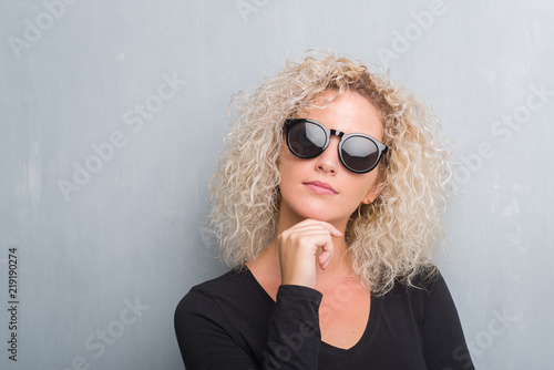 Young blonde woman with curly hair over grunge grey background with hand on chin thinking about question, pensive expression. Smiling with thoughtful face. Doubt concept.