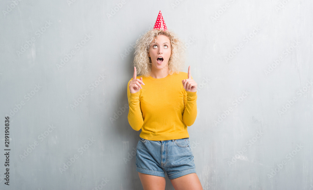 Young blonde woman over grunge grey wall wearing birthday hat amazed and surprised looking up and pointing with fingers and raised arms. Young blonde woman over grunge grey wall wearing birthday hat amazed and surprised looking up and pointing with fingers and raised arms.