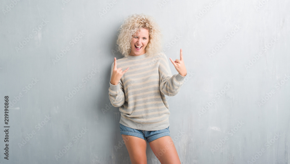 Young blonde woman with curly hair over grunge grey background shouting with crazy expression doing rock symbol with hands up. Music star. Heavy concept. Young blonde woman with curly hair over grunge grey background shouting with crazy expression doing rock symbol with hands up. Music star. Heavy concept.