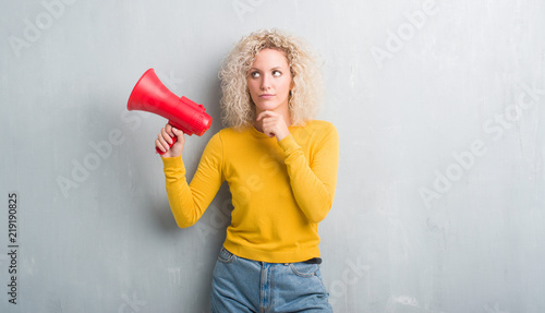 Young blonde woman over grunge grey background holding megaphone serious face thinking about question, very confused idea