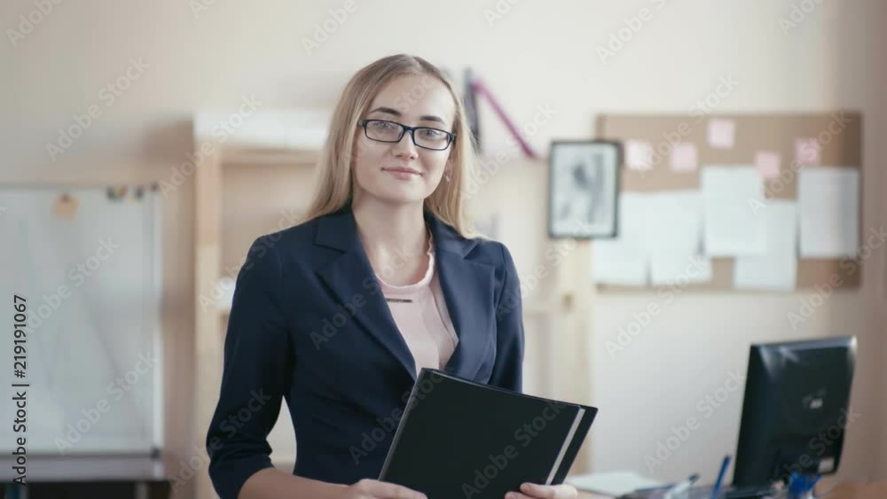 Call center employee at work in the office. A young girl with blond hair with glasses stands in the office, holding a folder for papers. The girl looks at the camera and smiles, laughs. Call center employee at work in the office. A young girl with blond hair with glasses stands in the office, holding a folder for papers. The girl looks at the camera and smiles, laughs.