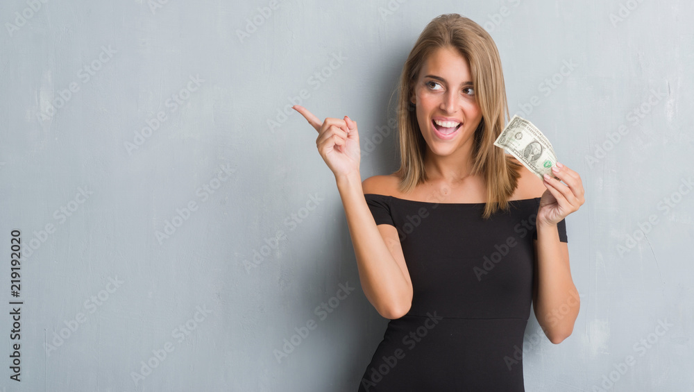 Beautiful young woman over grunge grey wall holding a dollar very happy pointing with hand and finger to the side Beautiful young woman over grunge grey wall holding a dollar very happy pointing with hand and finger to the side