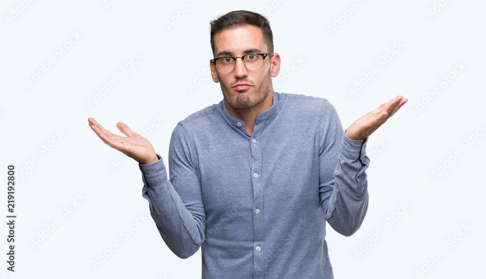 Fototapeta premium Handsome young elegant man wearing glasses clueless and confused expression with arms and hands raised. Doubt concept.