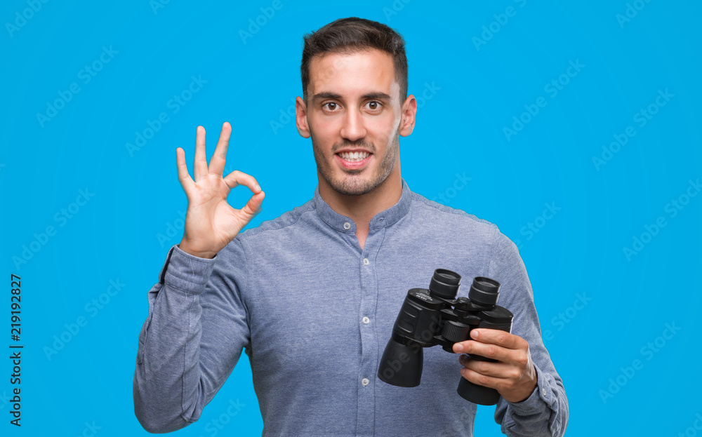 Handsome young man looking through binoculars doing ok sign with fingers, excellent symbol Handsome young man looking through binoculars doing ok sign with fingers, excellent symbol