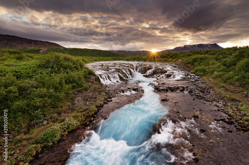 Large waterfall in a wide landscape with evening light  sunset with rays on a mountain range  water movement in long exposure - Location  Iceland  Golden circle  Bruarfoss