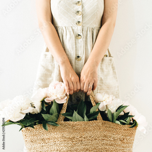 Young pretty woman in linen dress holding straw bag with white peony flowers on white background.