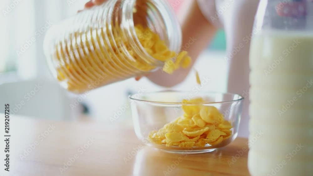 Woman hands pouring corn flakes into glass bowl. Close up preparing cereals with milk on kitchen table. Preparing traditional breakfast at home. Woman cooking on kitchen Woman hands pouring corn flakes into glass bowl. Close up preparing cereals with milk on kitchen table. Preparing traditional breakfast at home. Woman cooking on kitchen