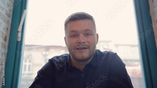A man speaks in a video chat sitting on the background of a window in a black shirt. Work on the Internet on a laptop