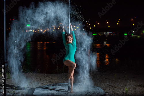 Red-haired woman in green body dancing on the pole and throwing flour against the city's city skyline. A young woman dances a striptease and throws up her hands a white powder