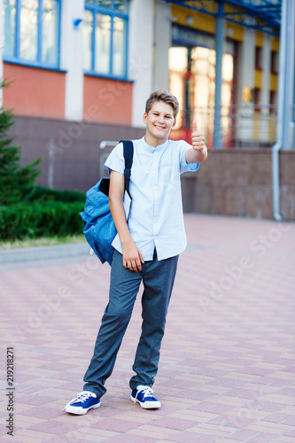 cute, young boy in blue shirt stands  in front of school with blue backpack and thumb up. Education, back to school concept © Natali