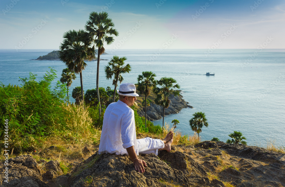 The man  in a white suit and hat sitting on a rock on the sea background