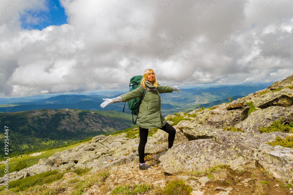 the blond tourist girl in a green jacket and with a backpack standing on a mountain resting and smiling, eyes closed and arms outstretched,against of mountains, forests and blue sky with white clouds the blond tourist girl in a green jacket and with a backpack standing on a mountain resting and smiling, eyes closed and arms outstretched,against of mountains, forests and blue sky with white clouds