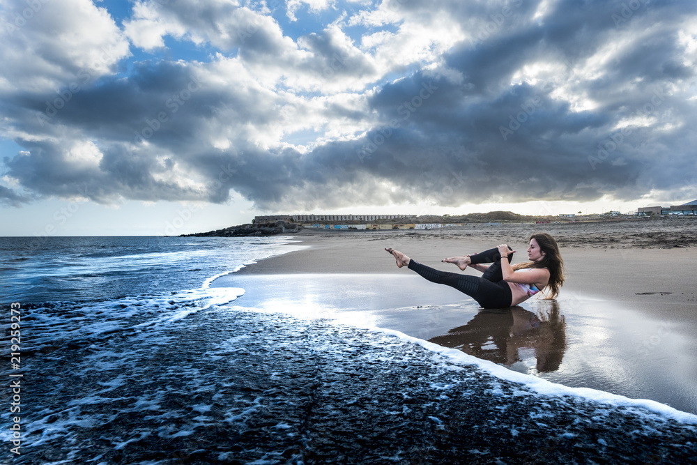 Naklejka premium blue colors epic style image with pretty brunette doing sport and pilates activity at the beach on the shore. scenic wave and clouds sunset on the background. balance lay down position for nice woman