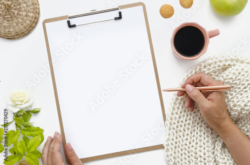 Clipboard Mock up.Top view hand of a woman holding a golden pen. Flat lay working table with a coffee, white rose, knitted scarf and apple. The girl makes a plan, fills document. Mockup for your idea photo