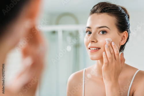 selective focus of smiling young woman applying face cream and looking at mirror in bathroom